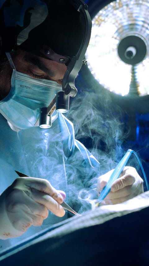 Cirurgião plástico realizando procedimento em centro cirúrgico com iluminação técnica e equipe médica.