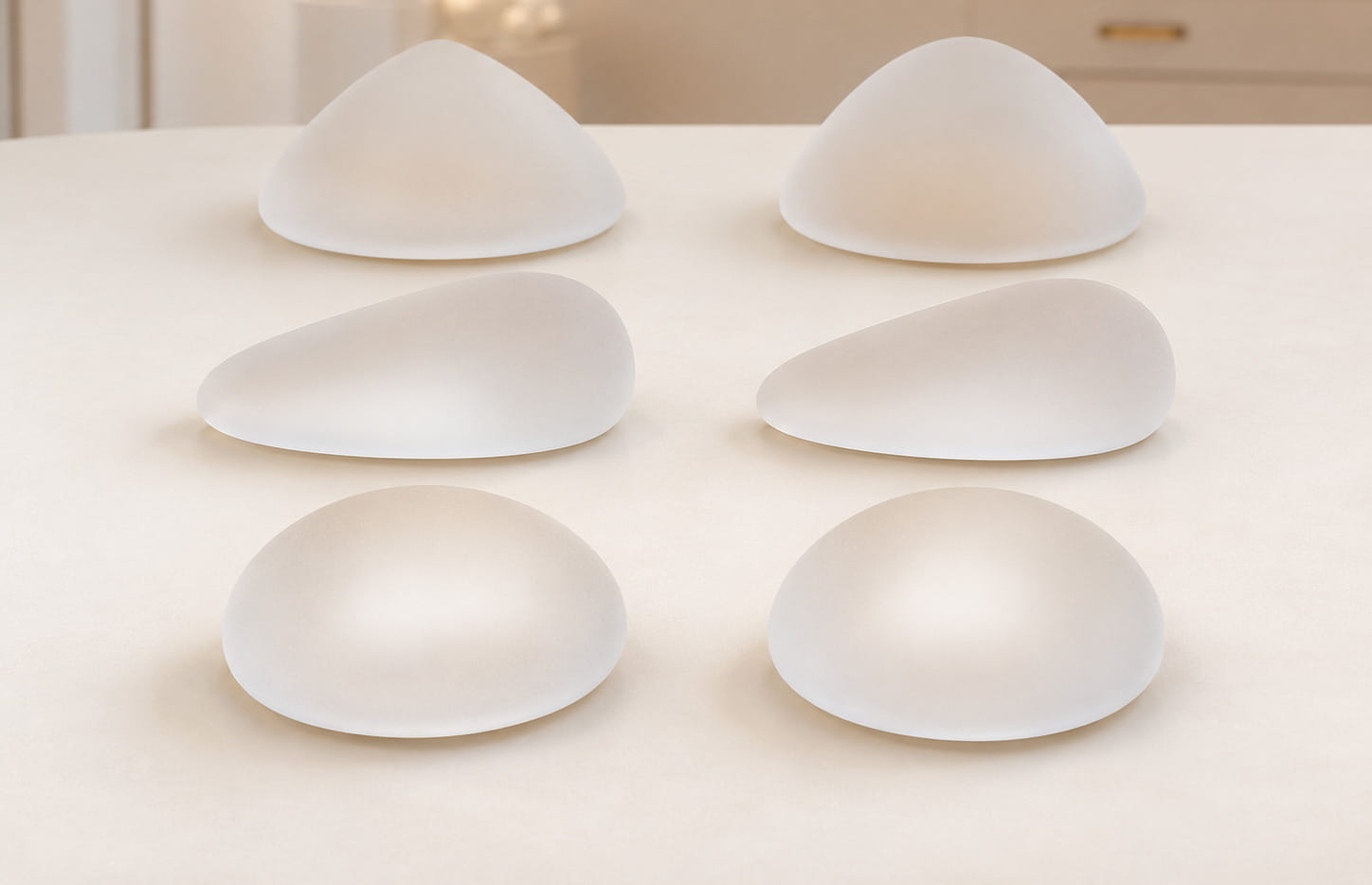 Modelos de prótese de silicone redonda, anatômica e cônica dispostos sobre mesa clara.